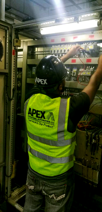 Onsite field service technician from Apex Industrial Automation working inside an industrial panel, wearing an Apex-branded safety helmet and high-visibility vest.