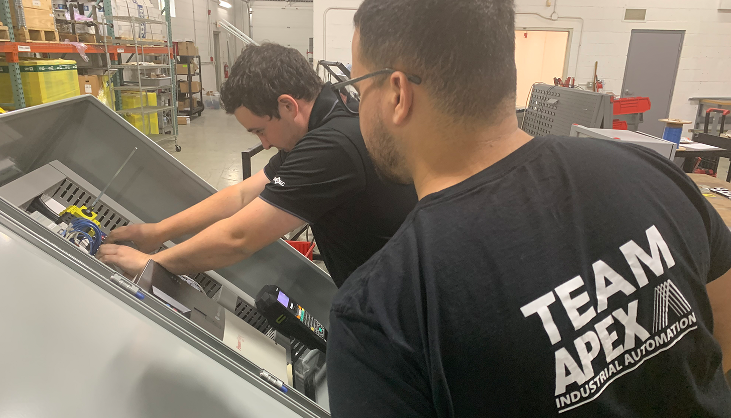 Two design engineers from Apex Industrial Automation fabricating a control panel, with one engineer reaching into the panel and another prominently featuring the Apex logo on his shirt.