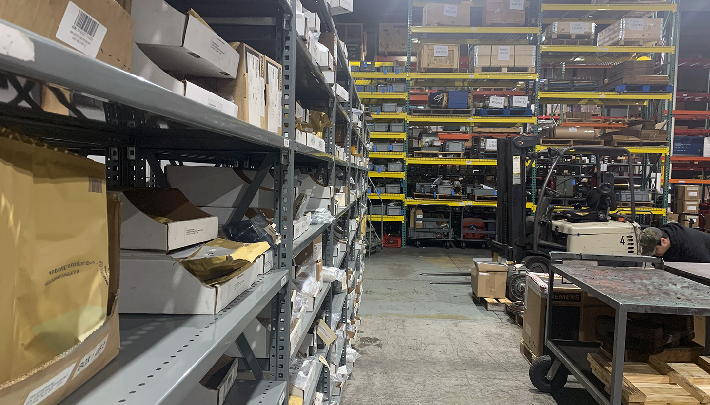 Interior view of Apex Industrial Automation's distribution warehouse showing industrial components on angled shelves, with large machinery in the background and a forklift on the right.