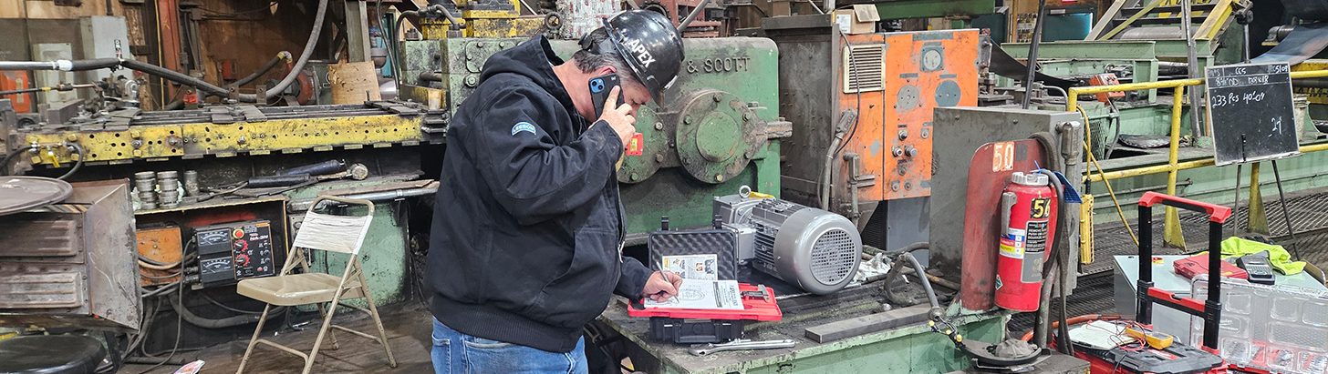Apex Industrial Automation onsite engineer taking notes on a phone at a customer site with old, messy industrial machinery in the background.
