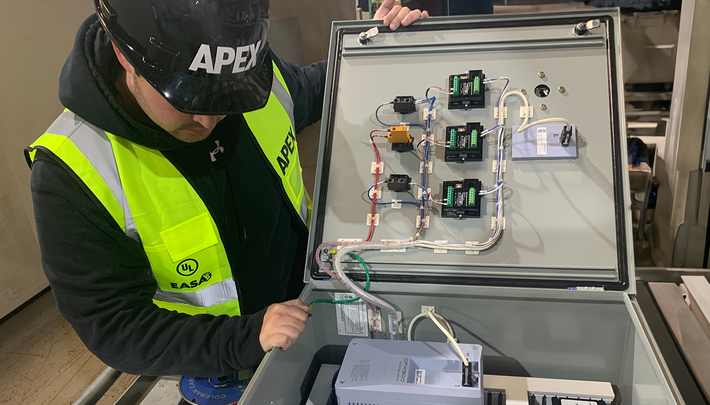 Apex Industrial Automation design engineer in branded safety vest and helmet inspecting an open control panel while doing onsite field service design at a customer site.