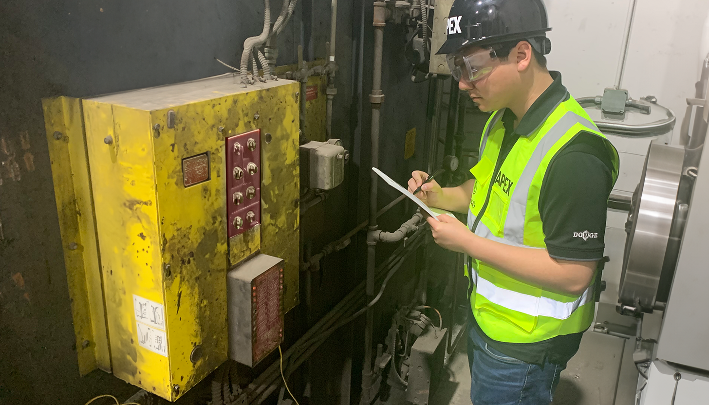 Apex Industrial Automation's onsite field service design engineer in branded safety vest and helmet surveying a large industrial machinery control panel.