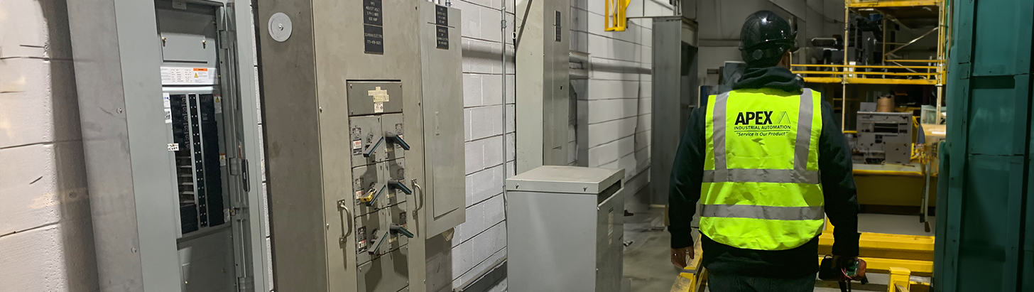 Apex Industrial Automation onsite engineer in branded safety vest and helmet walking through a customer's manufacturing plant, with wall panels and distant machinery visible.